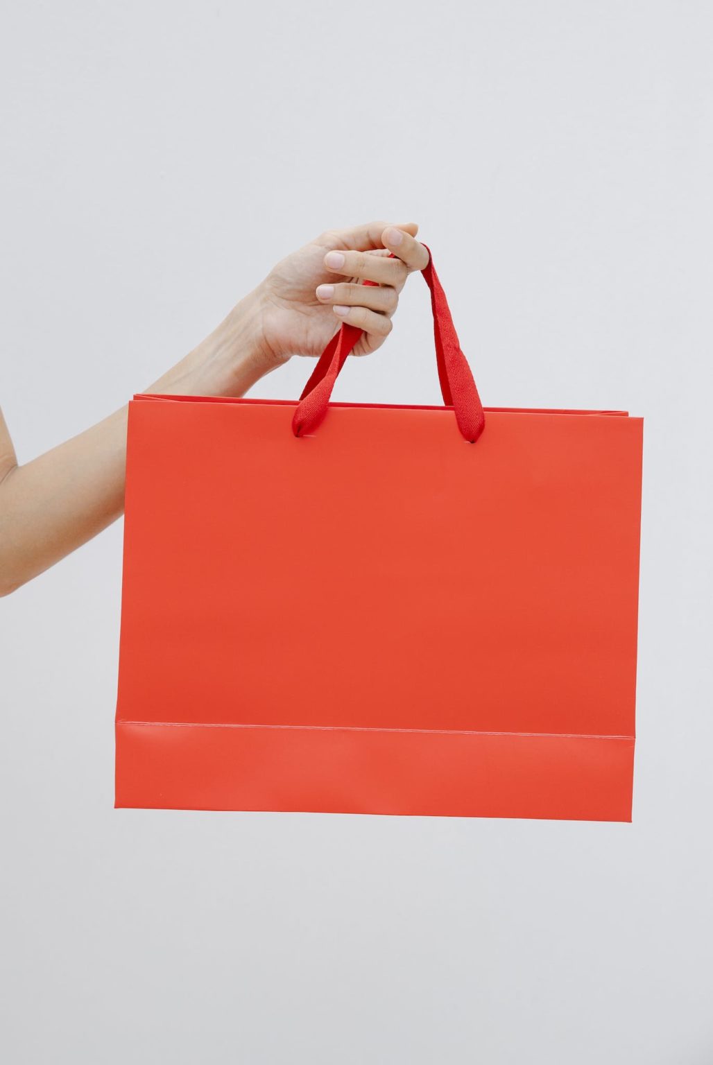 Crop unrecognizable female showing red paper shopping bag in hand against white wall
